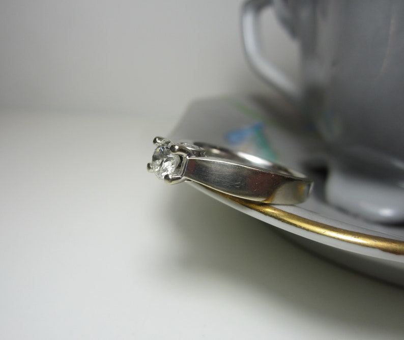 May include: A silver ring with a single round diamond set in the center. The ring is resting on a white saucer with a gold rim.