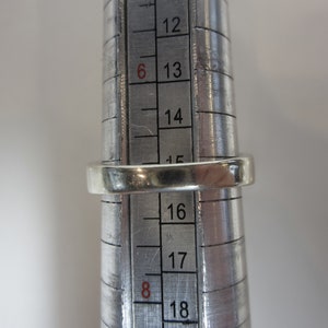 May include: A silver ring being sized on a metal ring sizer. The ring sizer shows sizes 12 through 18.
