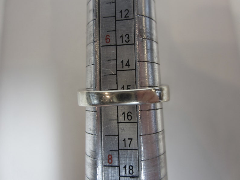 May include: A silver ring being sized on a metal ring sizer. The ring sizer shows sizes 12 through 18.