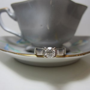 May include: A silver ring with a single round diamond set in the center. The ring is resting on a white saucer with a gold rim and blue floral pattern. The saucer is under a white teacup.
