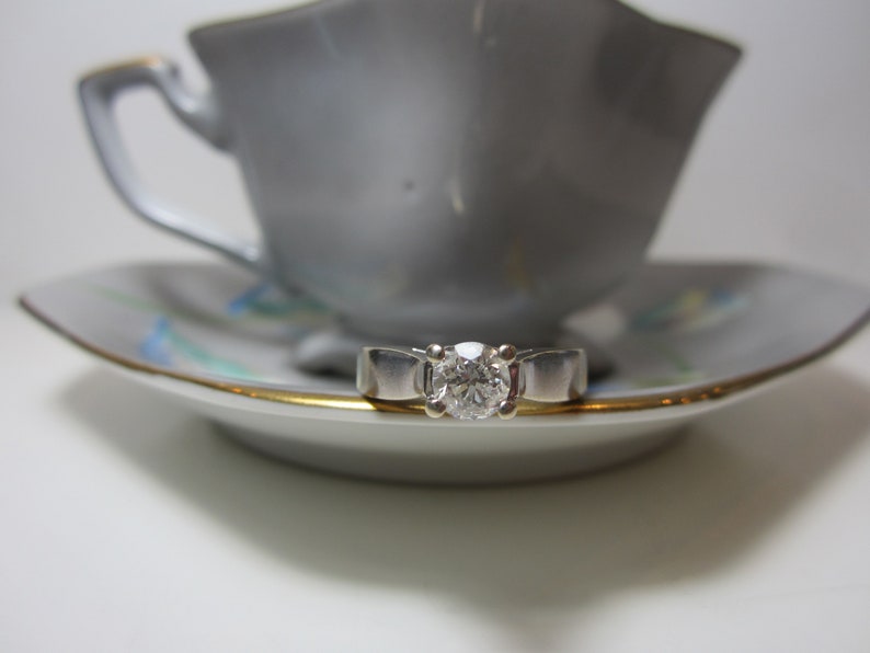May include: A silver ring with a single round diamond set in the center. The ring is resting on a white saucer with a gold rim and blue floral pattern. The saucer is under a white teacup.