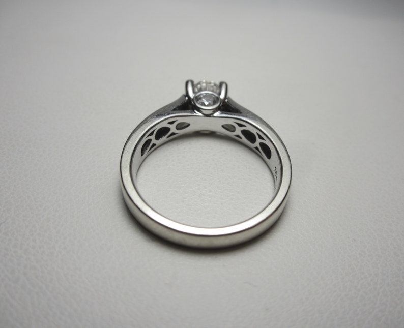 May include: A silver engagement ring with a single round diamond set in a prong setting. The band is decorated with a delicate filigree design.