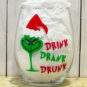 May include: A clear glass wine glass with a green Grinch face wearing a red Santa hat. The text "DRINK DRANK DRUNK" is printed on the glass in red.