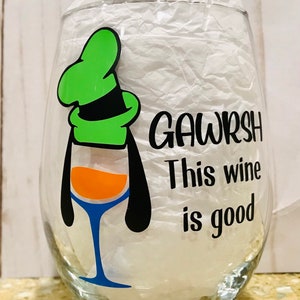 May include: A clear glass wine glass with a black and green cartoon character holding a glass of orange wine. The text "GAWRSH This wine is good" is printed on the glass.