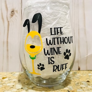 May include: A clear glass wine glass with a yellow and black cartoon dog design. The text "Life Without Wine Is Ruff" is printed on the glass.