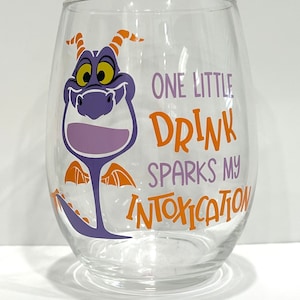 May include: A clear glass wine glass with a purple dragon cartoon character on the front. The dragon has orange horns and wings. The text on the glass reads "One little drink sparks my intoxication."