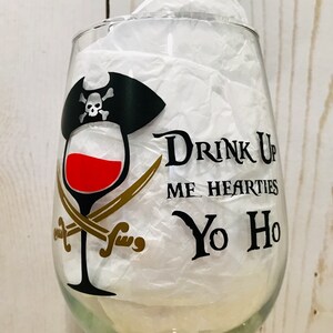 May include: A clear glass wine glass with a black and gold pirate design. The design includes a skull and crossbones, a pirate hat, crossed swords, and the text "Drink Up Me Hearties Yo Ho".