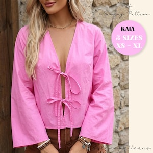 Può includere: Un top rosa acceso a maniche lunghe con scollatura a V profonda e chiusure con fiocco. Il top presenta una vestibilità ampia ed è realizzato in un tessuto leggero. L'immagine include anche un cerchio rosa con il testo "KAIA" e "5 TAGLIE XS-XL".