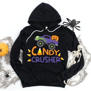 May include: Black hoodie with a purple monster truck carrying a jack-o'-lantern. The text "Candy Crusher" is written in orange and purple with candy corn accents.