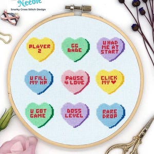 May include: A cross-stitch embroidery hoop with nine heart-shaped candies, each with a gaming phrase such as "U Fill My HP" and "Rare Drop." The design is set on light blue fabric, with the text "Notorious Needle" at the top. The hoop is approximately 20 cm in diameter.