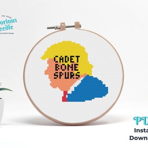 Puede incluir: Patrón de punto de cruz de un retrato de dibujos animados de una persona con cabello rubio y un traje azul, con el texto "CADET BONE SPURS" en letras negras. El patrón se muestra sobre un fondo blanco en un aro de bordado de madera.