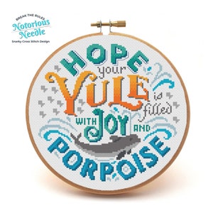 May include: A cross stitch hoop with a white background and colorful text that reads "Hope your Yule is filled with joy and purpose". The text is surrounded by a blue and green floral design. A gray dolphin is swimming in the bottom right corner of the hoop.
