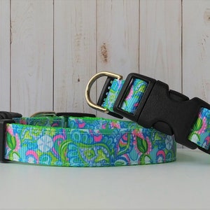 May include: A dog collar with a colorful floral pattern in shades of blue, green, and pink. The collar has a black plastic buckle and a gold-colored metal D-ring. The collar is made of a durable fabric.