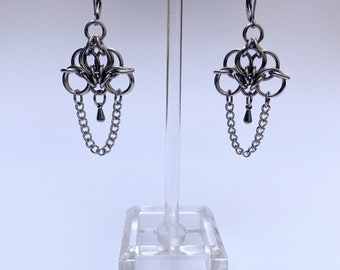 May include: A pair of silver earrings with a geometric design and a dangling chain. The earrings are displayed on a clear stand.