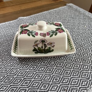 Op de afbeelding: Een witte keramische botervloot met deksel, versierd met bloemen- en vlindermotieven in roze, groen en paars. De rechthoekige schaal heeft een ronde knop op het deksel. De basis heeft een bijpassende bloemenrand.