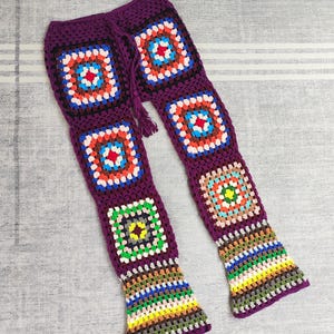 May include: Handmade crochet trousers in a deep purple shade, adorned with colourful square motifs. The squares incorporate red, blue, orange, and white elements. The trousers have a drawstring waist and flared bottoms, reflecting a bohemian aesthetic.
