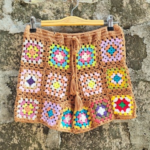 Könnte beinhalten: Braune Häkelshorts mit bunten quadratischen Mustern. Die Shorts hängen an einem Holzbügel. Die Quadrate sind in verschiedenen Farben, darunter Rosa, Gelb, Blau und Grün. Die Shorts haben einen Kordelzug und Quasten.