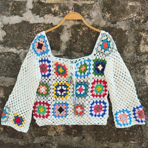 Può includere: Un top all'uncinetto bianco con scollatura quadrata e maniche lunghe. Il top presenta colorati quadrati della nonna in vari colori, tra cui blu, rosa, giallo e verde. Il top è appeso a una gruccia di legno.