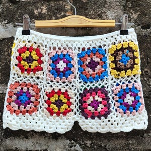 Può includere: Pantaloncini all'uncinetto bianchi con colorati quadrati della nonna nei toni del rosso, giallo, blu, viola e arancione. I pantaloncini sono esposti su una gruccia di legno su uno sfondo strutturato.