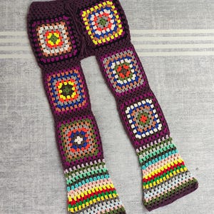 May include: Crocheted bell bottom pants with a colorful granny square pattern. The pants are made with a dark purple yarn and feature a variety of colors including red, yellow, blue, green, and orange.