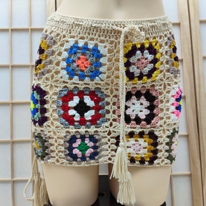 Puede incluir: Minifalda de crochet beige con patrones cuadrados coloridos. La falda tiene una cintura con cordón y borlas. Los cuadrados presentan colores vibrantes, incluyendo rojo, azul, amarillo y rosa.