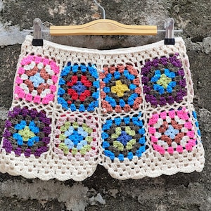 Peut inclure: Une jupe en crochet blanche avec un motif de carrés de grand-mère colorés. La jupe a un ourlet festonné et est faite de plusieurs couleurs, dont le rose, le bleu, le vert et le marron.