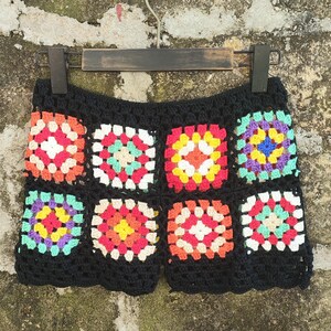 May include: Black crochet shorts with colorful granny square pattern. The squares feature red, orange, yellow, green, blue, and white. The shorts are hanging on a wooden hanger.