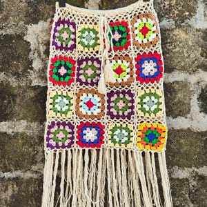 Op de afbeelding: Een handgemaakte, crèmekleurige gehaakte rok met een kleurrijk patchwork-ontwerp. De rok heeft vierkante motieven in verschillende kleuren, waaronder rood, groen, paars en geel. De rok heeft een gefranjerde zoom en een taille met trekkoord.