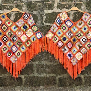 May include: A colorful, handmade crochet poncho with a V-neck design. The poncho features a patchwork pattern of squares in various colors, including orange, blue, yellow, and white. It has long orange fringe along the bottom edge, and is displayed on a wooden hanger.