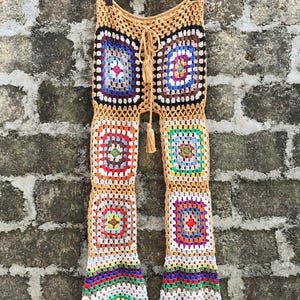 May include: Handmade crochet pants with a patchwork design. The pants feature a beige waistband and legs composed of colorful squares in various hues, including blue, red, green, and yellow. The pants have a drawstring closure and are displayed against a textured wall.