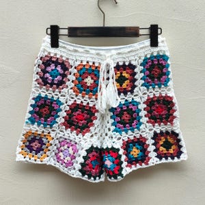 May include: Crochet shorts featuring a multi-colored granny square pattern with a white base. The shorts have a white drawstring waist with two tassels and are constructed from individual square motifs in shades of red, blue, yellow, and purple.