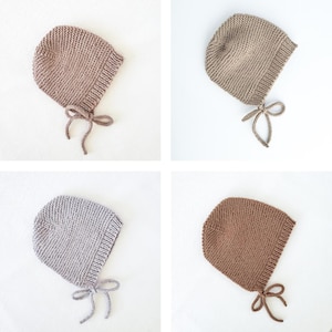 May include: Four knitted baby bonnets in different colors: beige, light brown, gray, and brown. The bonnets have a pointed top and a tie at the back.