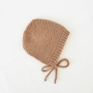 May include: A knitted bonnet in a warm, neutral brown shade. The bonnet has a rounded crown and a ribbed edge. A delicate bow is tied under the chin. The soft, textured knit suggests a cosy, handmade item, perfect for a baby.