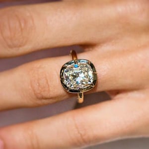 May include: A gold ring featuring a large, cushion-cut center stone with a faceted surface. The stone is set in a metallic bezel setting that frames the gem, mounted on a thin, polished gold band that encircles the finger.