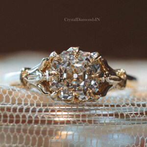May include: A gold ring featuring a central cushion-cut clear gemstone held by multiple prongs. The band displays an intricate, ornate metalwork design with decorative filigree patterns extending from the setting. The text CrystalDiamondsIN is visible.