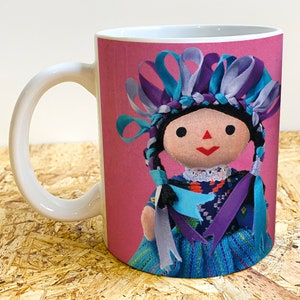 Puede incluir: Taza de cerámica blanca con fondo rosa y una muñeca colorida con cintas azules, moradas y grises en el pelo. La muñeca lleva un vestido azul y morado.