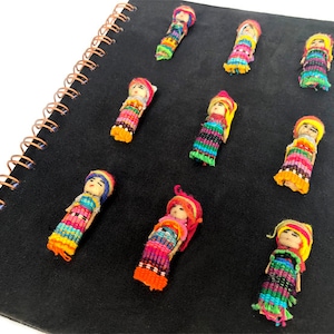 Puede incluir: Una colección de pequeñas muñecas hechas a mano con ropa tejida colorida y cabello de hilo. Las muñecas están dispuestas sobre una superficie negra, posiblemente un cuaderno, con una encuadernación en espiral de color cobre. Los atuendos de las muñecas presentan tonos vibrantes de rojo, amarillo, azul y verde.
