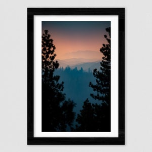 May include: A photograph of a mountain range in the distance, framed by two tall, dark trees. The sky is a soft pink and orange, with a hazy blue layer in the foreground.
