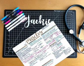 May include: A black cutting mat with a white grid pattern and a stethoscope on top. The mat has a white label that says "Vintage Cutting Mat 18" x 12". There are also several pens and markers on the mat. A sheet of paper with handwritten notes about muscles is on the mat.