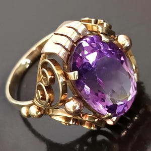 Könnte beinhalten: Ein Goldring mit einem großen, oval geschliffenen violetten Edelstein in einer dekorativen Metallfassung. Die Fassung zeichnet sich durch filigrane Schnörkel und kleine kugelförmige Akzente aus, die den zentralen Stein umgeben.