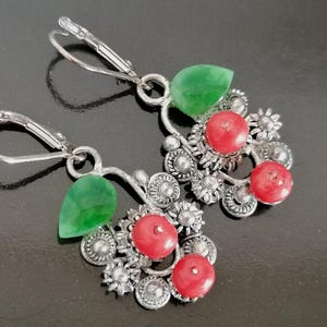 May include: Silver dangle earrings with red and green gemstone accents. The earrings feature a cluster of red gemstones and a single green gemstone on each earring.