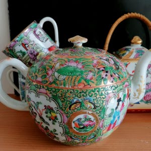 May include: A porcelain tea set featuring a teapot, a teacup, and a tea caddy. The set is decorated with colorful floral and bird motifs, with gold accents. The teapot has a rounded shape and a curved spout. The tea caddy has a woven handle.