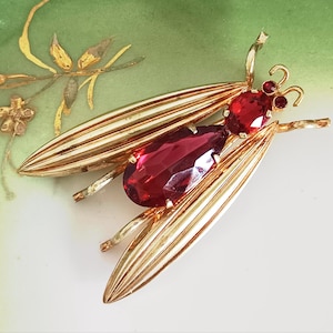 May include: A gold-toned insect brooch featuring long, ribbed wings and a body accented with a large, teardrop-shaped red gemstone and a smaller oval red gemstone. The head includes two small red circular stones and two thin antennae.