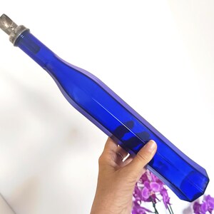 Op de afbeelding: Een lange, kobaltblauwe glazen fles met een gefacetteerd, zeshoekig lichaam en een smalle hals. De fles is voorzien van een zilverkleurige metalen schenktuit. Het glas is doorschijnend, waardoor licht door de diepblauwe kleur heen schijnt.