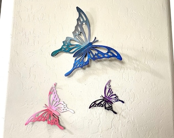 May include: Three metal butterfly wall decor pieces in different sizes and colors. The largest butterfly is blue and purple, the medium butterfly is pink and white, and the smallest butterfly is purple.