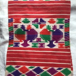May include: A white and red woven textile with a colorful geometric pattern. The pattern features red, green, purple, and white colors. The textile has a fringed edge.
