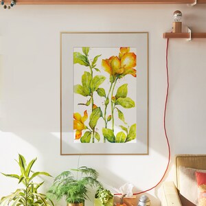 May include: A framed watercolor painting of yellow flowers with green leaves on a white background. The painting is hanging on a white wall above a wooden shelf with a small potted plant and a piece of driftwood. A couch with a throw pillow is in the foreground.