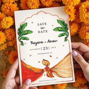 May include: A rectangular save the date card featuring a decorative arch outline, green banana leaf illustrations, and a tied orange and beige fabric graphic. Text reads: SAVE the DATE, Nayana & Aarav, NOVEMBER 23 20XX, POUGHKEEPSIE, NY, Formal Invitation to follow.