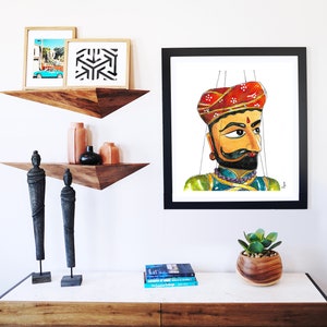 Puede incluir: Una pintura de acuarela enmarcada de un t&iacute;tere tradicional indio con un turbante rojo, un atuendo verde y dorado y un bigote. El t&iacute;tere est&aacute; suspendido por cuerdas y tiene una expresi&oacute;n seria.