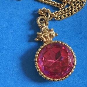 May include: Gold-toned necklace with a large, round, faceted red pendant. The pendant is set in a gold frame and hangs from a matching gold chain. The necklace is displayed on a blue background.